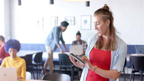 Young-Caucasian-woman-in-casual-business-attire-reviews-a-tablet-in-a-busy-office