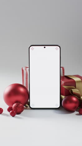 Vertical-video-of-christmas-red-baubles,-presents-and-smartphone-with-copy-space-on-white-background