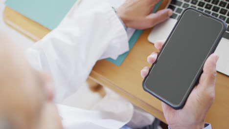 Senior-biracial-male-doctor-using-laptop-and-smartphone,-copy-space-on-screen