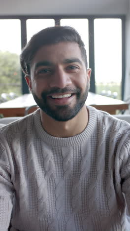 Vertical-video-of-portrait-of-happy-biracial-man-sitting-on-sofa-at-home,-slow-motion