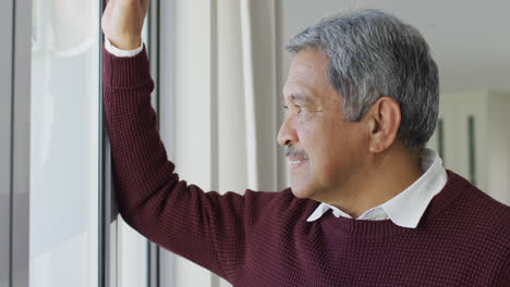 Video-of-thoughtful-senior-biracial-man-in-living-room-looking-out-of-window-and-smiling-to-camera