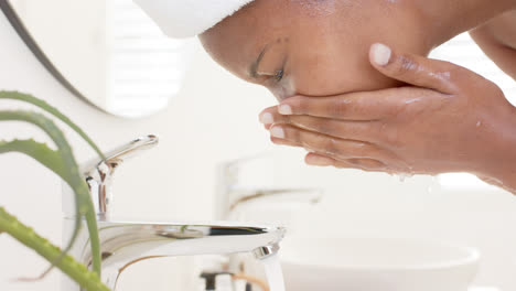 Biracial-woman-wearing-bathrobe-and-washing-her-face-in-front-of-mirror-in-bathroom,-slow-motion