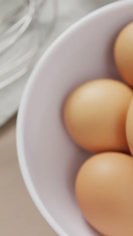 Video-of-overhead-view-of-white-bowl-with-eggs-and-egg-whisk-on-beige-background