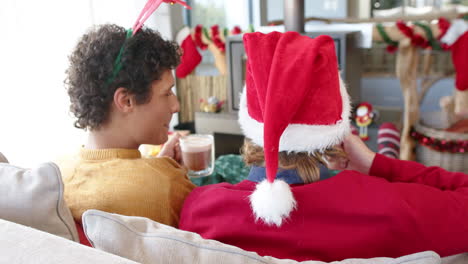 Diverse-gay-male-couple-drinking-cocoa-on-sofa-by-fireplace-at-home-at-christmas,-slow-motion