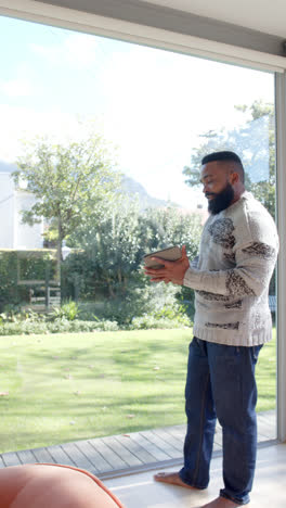 Video-Vertical-De-Un-Hombre-Afroamericano-Usando-Una-Tableta-Y-Mirando-Por-La-Ventana-En-Casa,-Cámara-Lenta