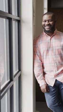 Retrato-En-Video-Vertical-De-Un-Feliz-Hombre-De-Negocios-Afroamericano-Casual-Parado-Junto-A-La-Ventana-En-La-Oficina