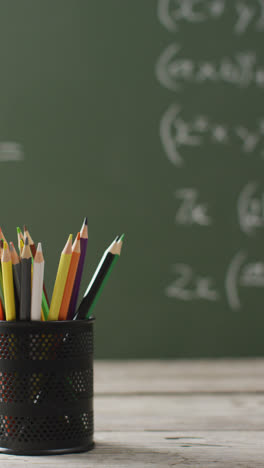 Vertical-video-of-coloured-pencils-in-pot-on-desk-with-mathematics-on-chalk-board-in-background