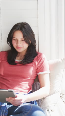 Vertical-video-of-happy-biracial-woman-sitting-on-bed-using-tablet-in-sunny-room,-slow-motion