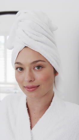 Vertical-video-of-portrait-of-happy-biracial-woman-with-towel-on-head-in-bathroom,-slow-motion