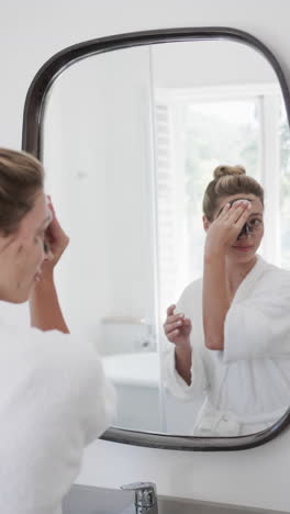 Vertical-video-of-biracial-woman-applying-beauty-face-mask-in-bathroom,-slow-motion