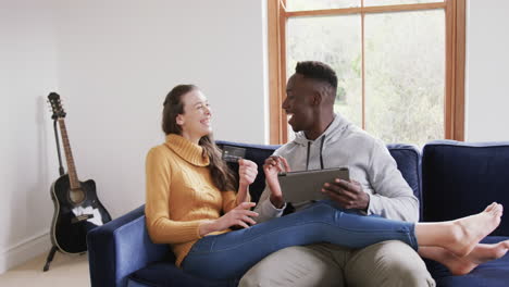 Happy-diverse-couple-sitting-on-sofa,-using-tablet-and-credit-card-in-home,copy-space