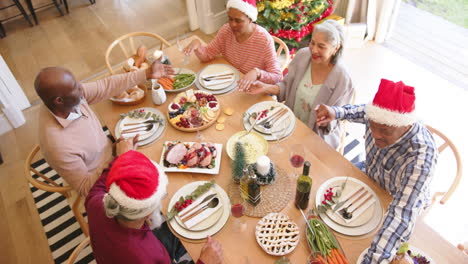 Felices-Y-Diversos-Amigos-Mayores-Tomados-De-La-Mano-Para-Dar-Las-Gracias-En-La-Mesa-Del-Comedor-Navideño,-En-Cámara-Lenta