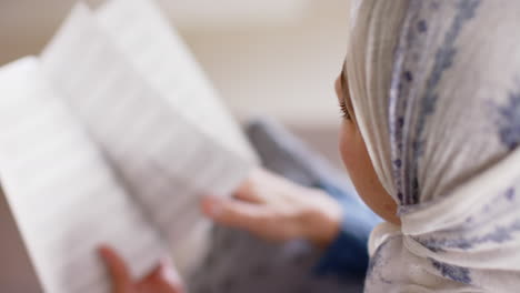 Happy-biracial-woman-in-hijab-reading-book-on-sofa-at-home-with-copy-space,-slow-motion