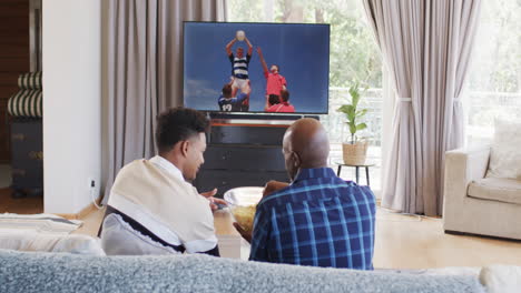 Vista-Trasera-De-Un-Padre-Afroamericano-Y-Un-Hijo-Adulto-Comiendo-Palomitas-De-Maíz-Viendo-Rugby-En-La-Televisión,-Cámara-Lenta