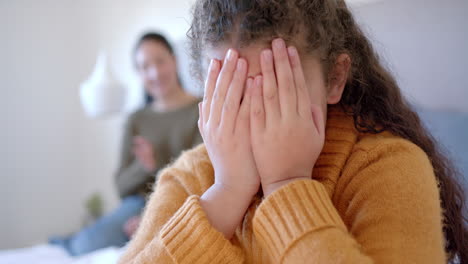 Nervous-biracial-mother-admonishing-daughter-on-bed-in-sunny-bedroom