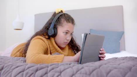 Happy-biracial-girl-lying-on-bed,-using-tablet-and-headphones-in-sunny-bedroom