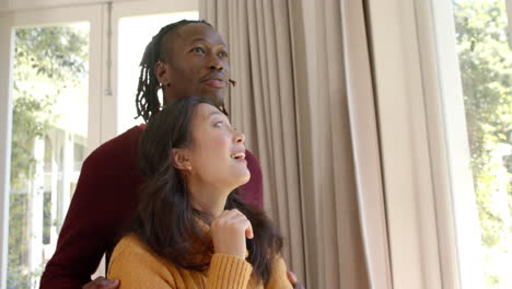 Happy-diverse-couple-embracing-and-looking-out-of-sunny-window-at-home,-copy-space,-slow-motion