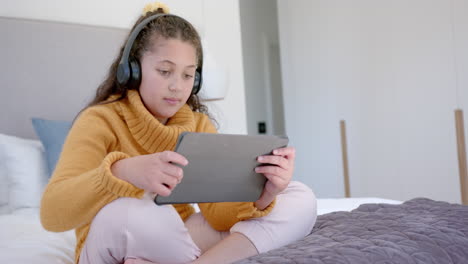 Happy-biracial-girl-sitting-on-bed,-using-tablet-and-headphones-in-sunny-bedroom