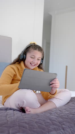 Vertical-video-of-happy-biracial-girl-sitting-on-bed,-using-tablet-and-headphones-in-sunny-bedroom