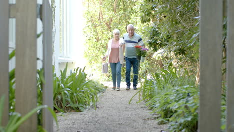 Älteres,-Kaukasisches-Paar,-Das-Blumen-Hält-Und-Im-Sonnigen-Garten-Spazieren-Geht,-Zeitlupe