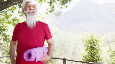 Feliz-Hombre-Mayor-Birracial-Con-Barba-Blanca-Sosteniendo-Estera-De-Yoga-Sonriendo-Al-Sol,-Espacio-Para-Copiar,-Cámara-Lenta