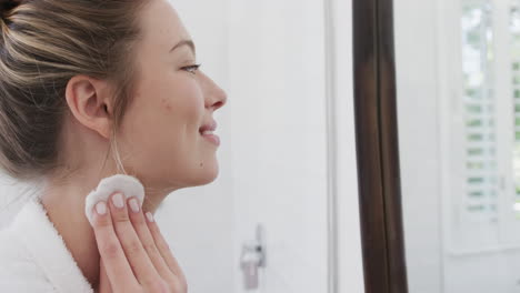 Happy-biracial-woman-cleansing-face-using-cotton-pad-looking-in-mirror-in-bathroom,-slow-motion