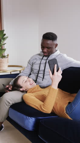 Happy-diverse-couple-sitting-and-lying-on-sofa-and-using-smartphone-in-sunny-living-room,-copy-space