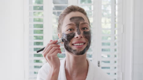 Portrait-of-happy-biracial-woman-applying-beauty-face-mask-in-bathroom,-slow-motion