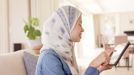 Happy-biracial-woman-in-hijab-using-tablet-on-sofa-at-home-with-copy-space,-slow-motion