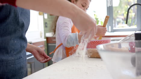 Happy-biracial-mother-and-daughter-preparing-dough-and-smiling-in-sunny-kitchen