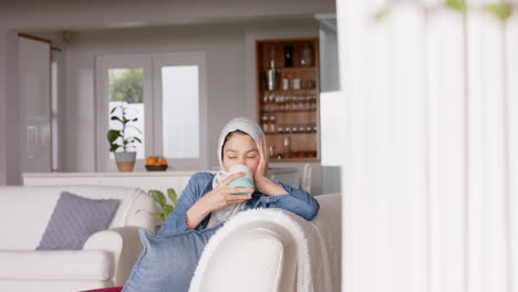 Happy-biracial-woman-in-hijab-drinking-tea-on-sofa-at-home-with-copy-space,-slow-motion