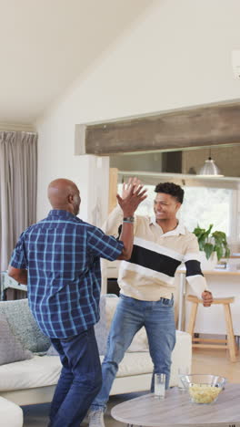 Vertical-video-of-african-american-father-and-son-cheering-sport-on-tv-and-high-fiving,-slow-motion