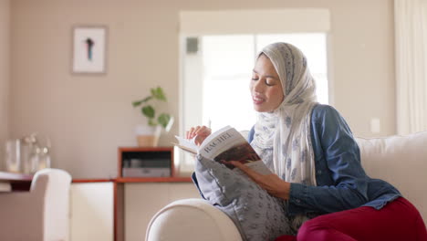 Feliz-Mujer-Birracial-En-Hijab-Leyendo-Un-Libro-En-Un-Sofá-En-Casa-Con-Espacio-Para-Copiar,-Cámara-Lenta