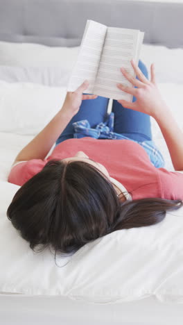 Vertical-video-of-biracial-woman-lying-on-bed-reading-book,-slow-motion