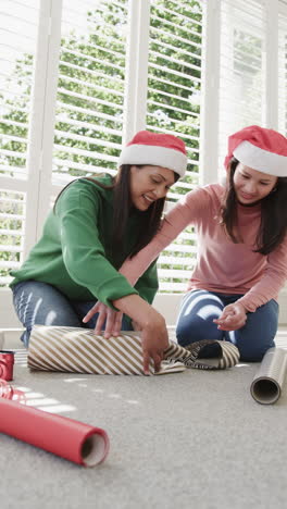 Vertical-video-of-happy-biracial-mother-and-daughter-in-christmas-hats-wrapping-gift,-slow-motion