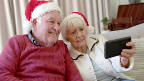 Pareja-Caucásica-Mayor-Con-Sombreros-De-Santa-Usando-Un-Teléfono-Inteligente-Para-Videollamadas-Navideñas,-Cámara-Lenta