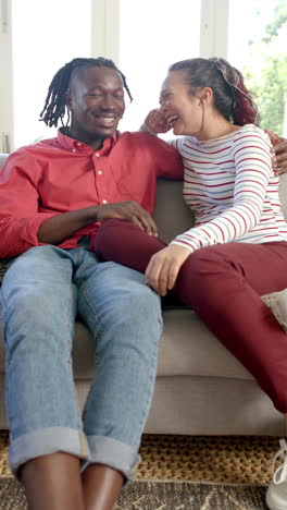 Vertical-video-portrait-of-happy-diverse-couple-sitting-on-couch-in-sunny-living-room,-slow-motion