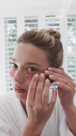 Vertical-video-of-biracial-woman-applying-under-eye-patches-in-bathroom,-slow-motion