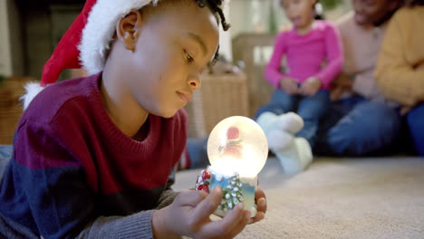 Afroamerikanischer-Sohn-In-Weihnachtsmütze-Hält-Leuchtende-Schneekugel,-Familie-Im-Hintergrund,-Zeitlupe