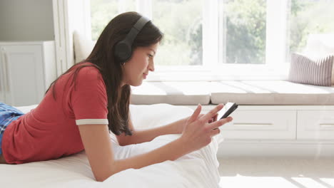 Mujer-Birracial-Feliz-Con-Auriculares-Acostada-En-La-Cama-En-Una-Habitación-Soleada-Usando-Tableta,-Espacio-Para-Copiar,-Cámara-Lenta