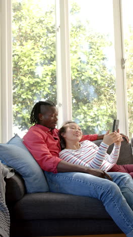 Vertical-video-of-happy-diverse-couple-using-tablet-in-sunny-living-room,-copy-space,-slow-motion