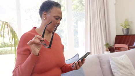 Feliz-Mujer-Afroamericana-Mayor-Con-Gafas-Usando-Un-Teléfono-Inteligente-En-Una-Sala-De-Estar-Soleada,-Cámara-Lenta