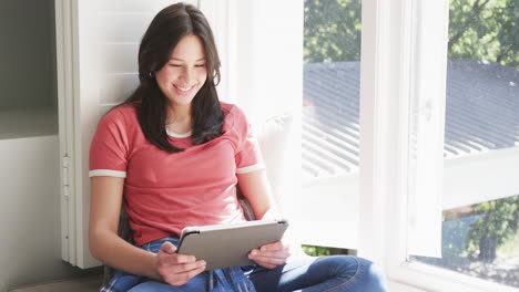 Feliz-Mujer-Birracial-Sentada-Junto-A-La-Ventana-En-Una-Habitación-Soleada-Usando-Tableta,-Espacio-Para-Copiar,-Cámara-Lenta