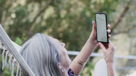 Caucasian-middle-aged-woman-using-smartphone-relaxing-in-hammock-on-terrace-in-nature,-slow-motion