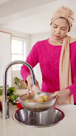 Vertical-video-of-biracial-woman-in-hijab-washing-food-in-kitchen-at-home,-copy-space,-slow-motion
