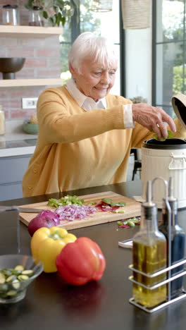 Vertical-video-of-senior-caucasian-woman-cleaning-vegetable-peels-in-kitchen-at-home,-slow-motion