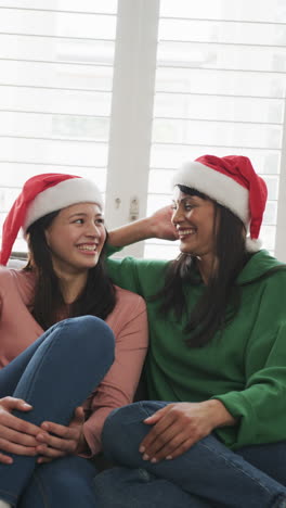 Vertical-video-of-happy-biracial-mother-and-daughter-in-christmas-hats-sitting-on-couch,-slow-motion