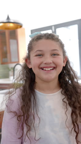 Vertical-video-of-happy-biracial-girl-with-long,-curly-hair-standing-and-smiling-in-sunny-kitchen