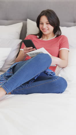 Vertical-video-of-happy-biracial-woman-sitting-on-bed-reading-book-and-using-smartphone,-slow-motion