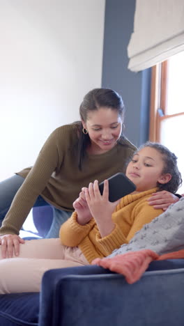 Feliz-Madre-E-Hija-Birracial-Sentadas-En-El-Sofá-Y-Usando-Un-Teléfono-Inteligente-En-Una-Soleada-Sala-De-Estar